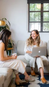 Deux jeunes femmes souriantes discutant, l’une tenant un MacBook, l’autre une tasse de café
