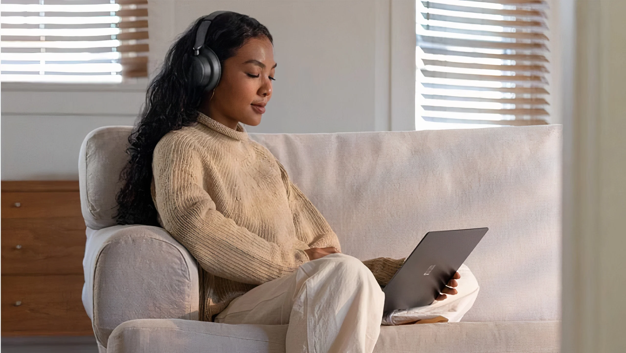 Jeune fille assise détendue sur un canapé, casque sur les oreilles, ordinateur portable sur les genoux, regard concentré sur l’écran