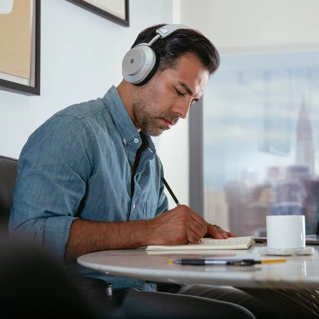 Homme brun portant un casque sur les oreilles, assis à une table, en train d’écrire