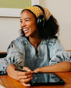 Jeune fille avec bandana jaune, casque sur les oreilles, tenant un stylet et une tablette, souriant à une personne à sa gauche, formation d’anglais interactive
