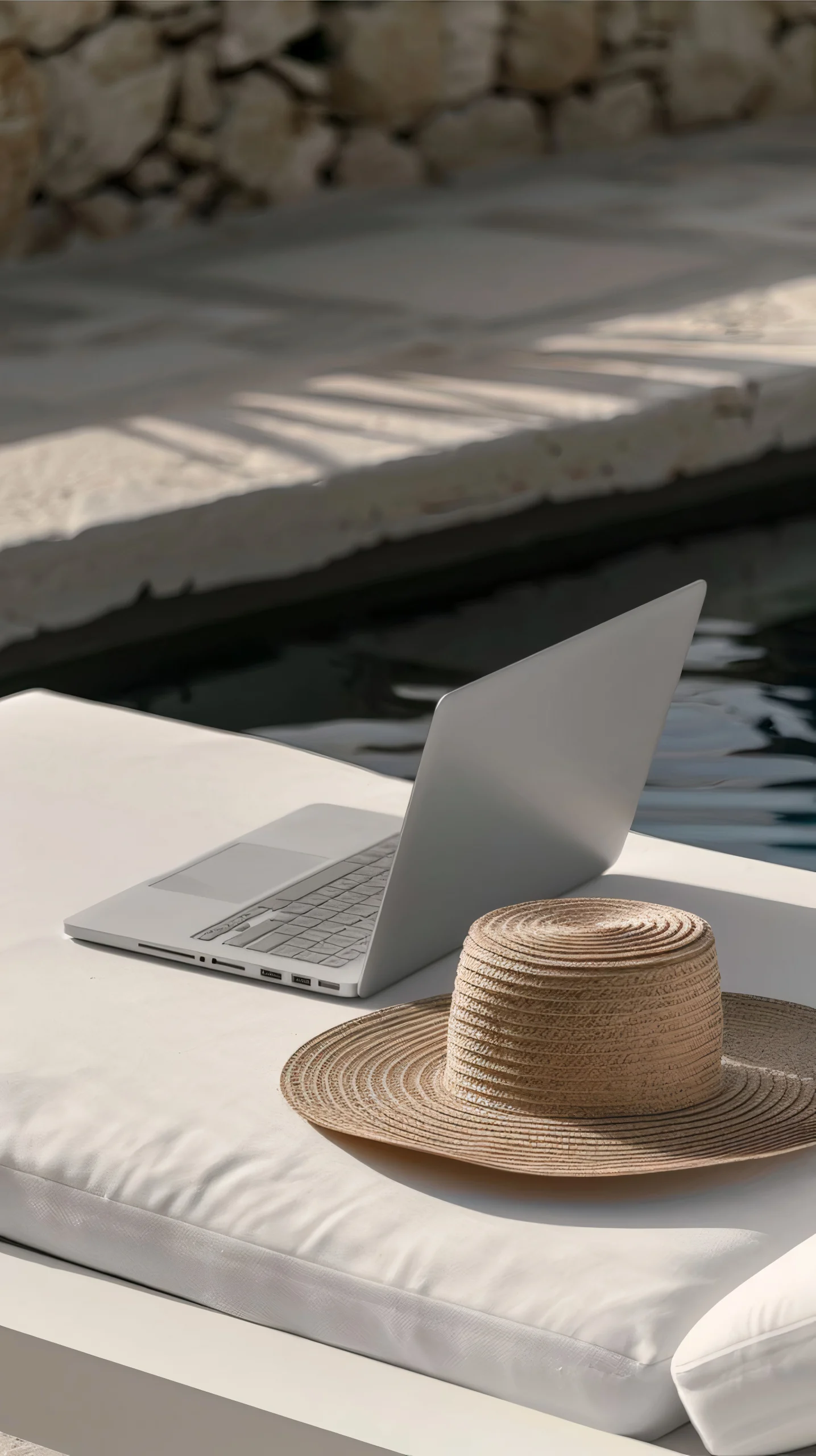 Ordinateur portable posé sur un transat à côté d’un chapeau de paille au bord d’une piscine