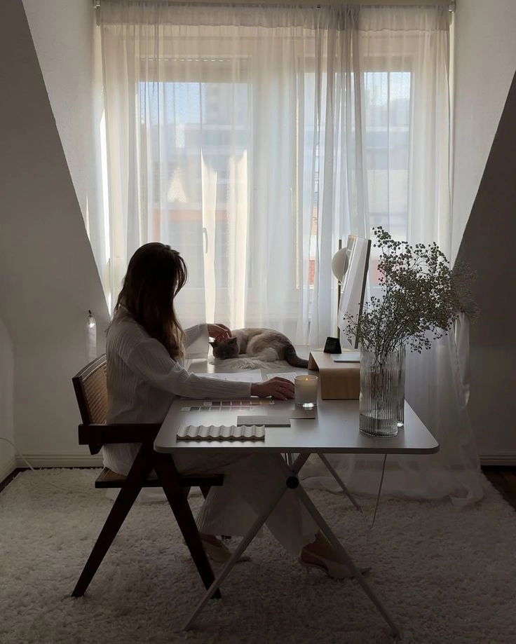 Femme assise à son bureau devant une fenêtre, caressant son chat posé sur le bureau